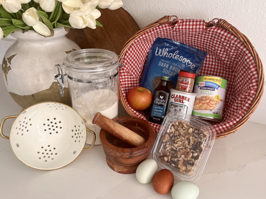 Ingredients for Apple Walnut Cake. Walnuts, canned apple pie filling, eggs, baking powder, flour, vanilla, brown sugar, pumpkin pie spice and an apple in a basket on the counter