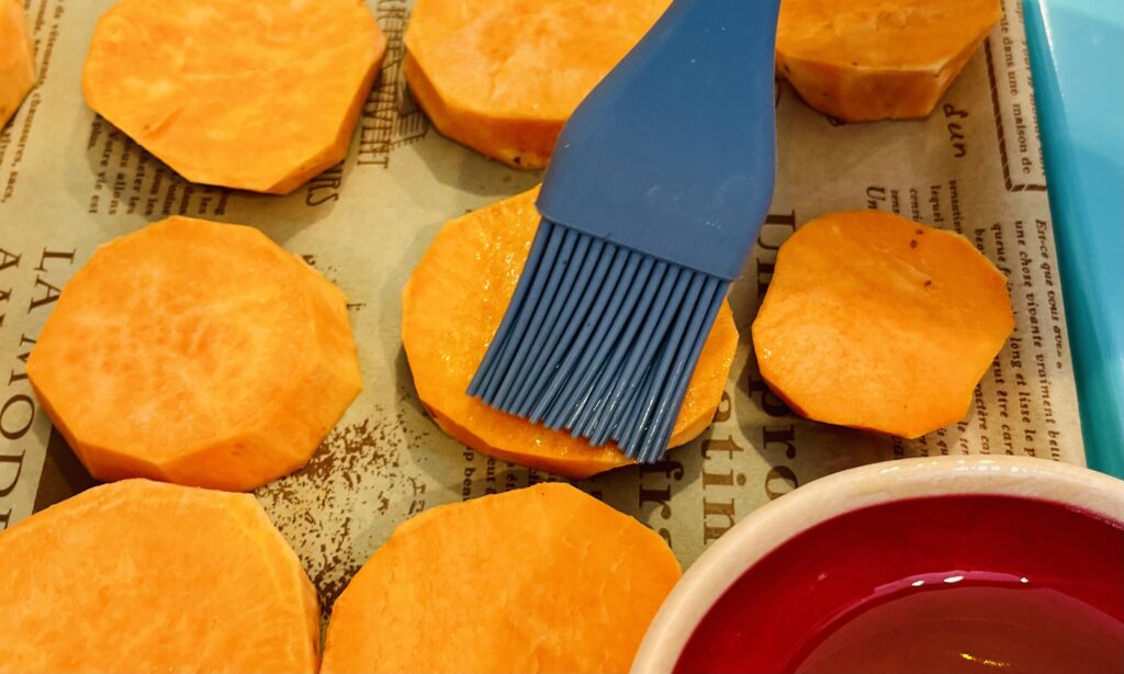 Sweet potato rounds being brushed with olive oil on a lined baking sheet
