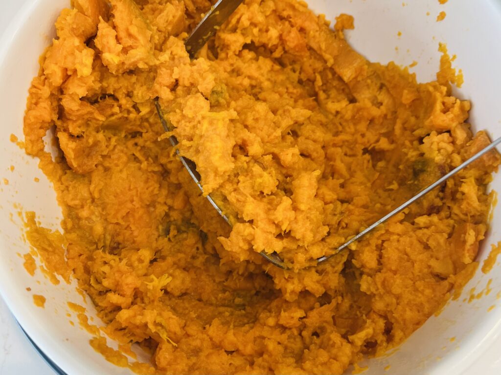 Mashing with a potato masher- sweet potatoes with cashew milk in a large bowl
