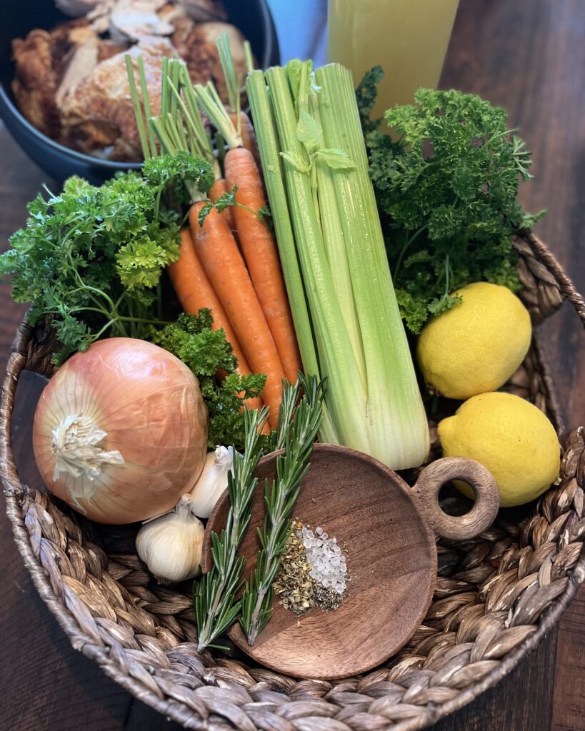 Ingredients -carrots, celery, parsley, onion, garlic, rosemary, lemon, store bought rotisserie chicken, salt, pepper, herb de Provence and chicken broth in a basket on the table