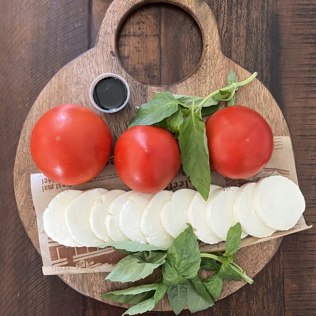 Caprese Salad ingredients, tomatoes, pre-sliced mozzarella cheese, bunch of basil, balsamic glaze on a cutting board