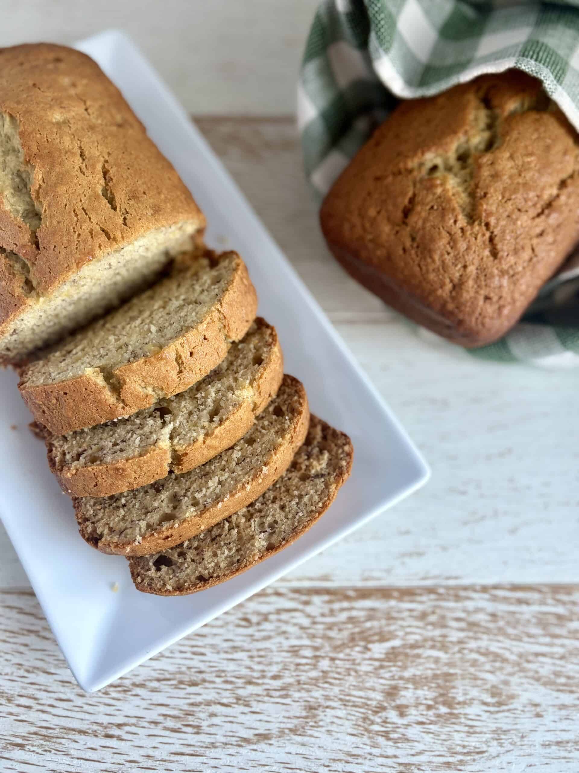 sliced banana bread on a white plate next to a second loaf wrapped in a kitchen towel