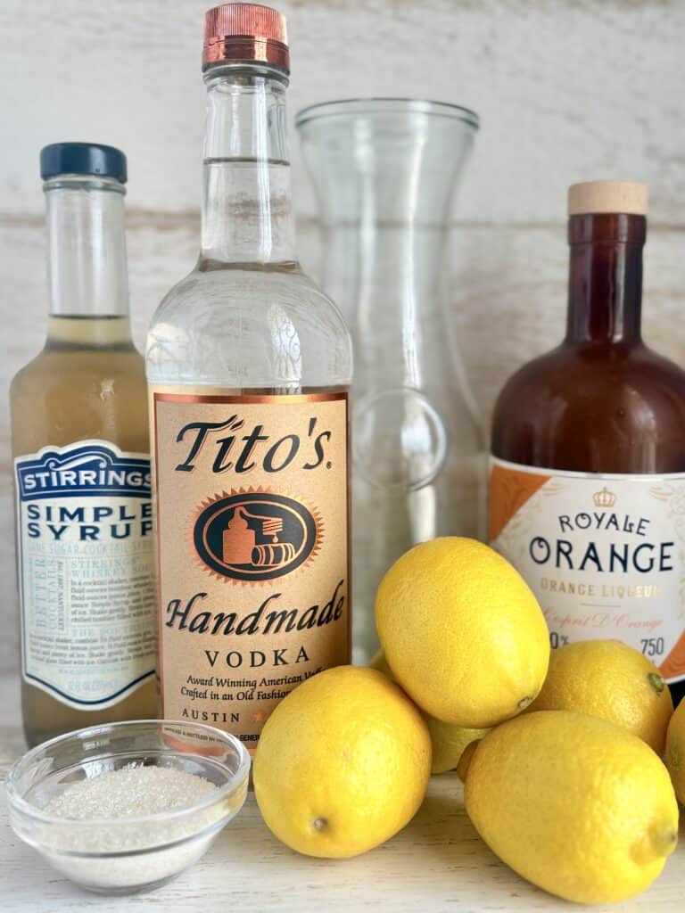 vodka, simple syrup, orange liqueur, lemons and small bowl with sugar on a wooden board.
