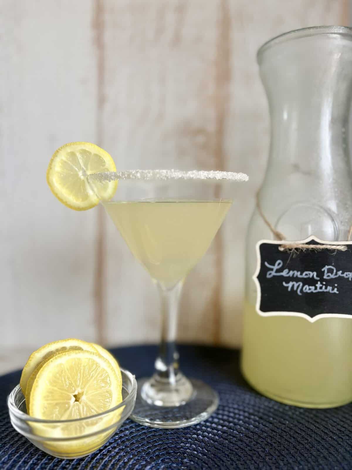 Elegant Lemon Drop Martini garnished with a lemon wheel and sugar-rimmed glass, served alongside a pitcher of fresh lemon cocktail. Perfect for a crowd, this easy pitcher cocktail recipe is ideal for.