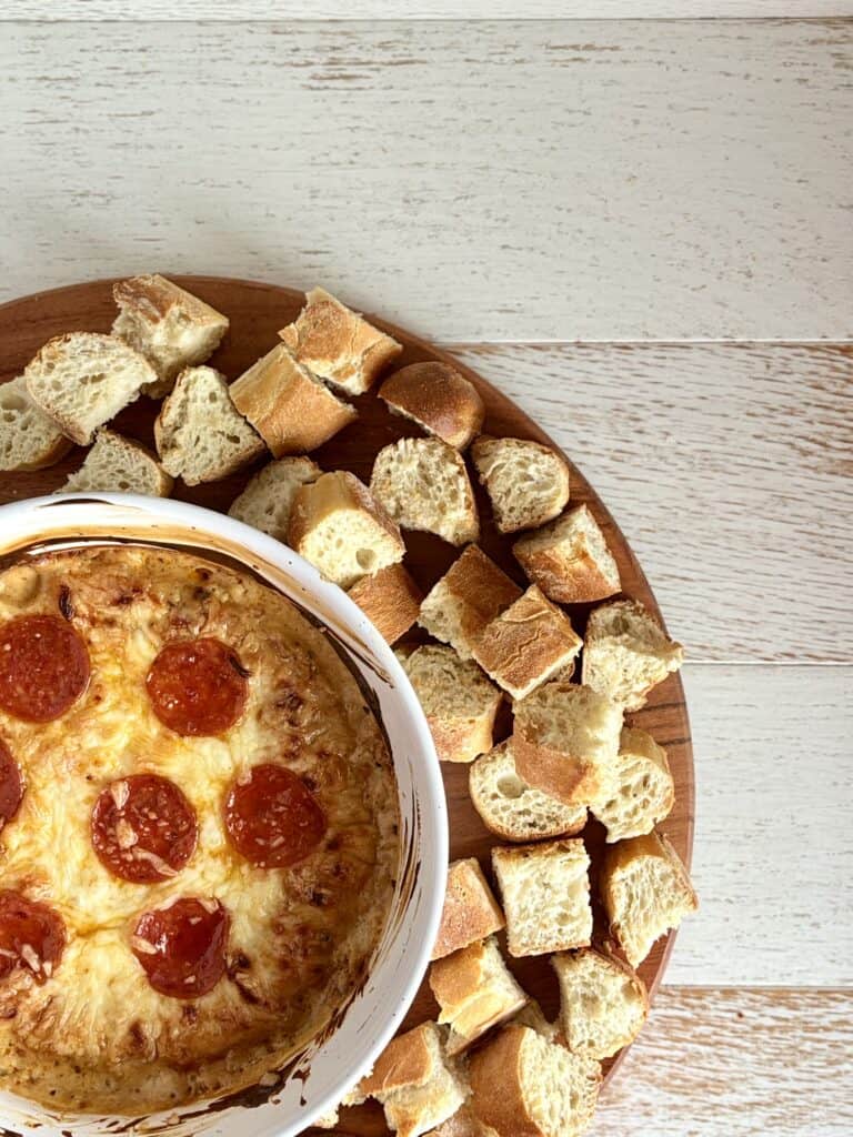Close-up of hot white pizza dip topped with pepperoni slices, served with cubed bread on a wooden platter.