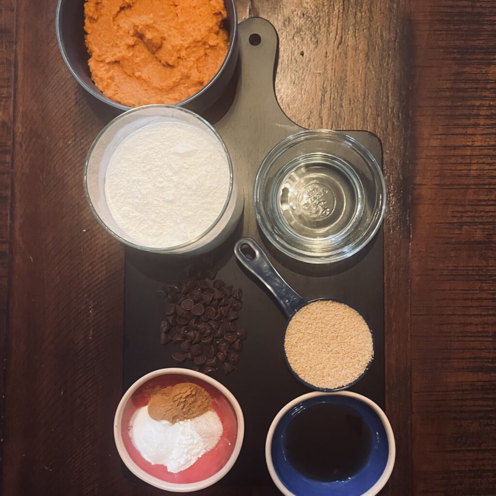 pumpkin puree, vegetable oil, brown sugar maple syrup, flour, pumpkin pie spice , baking powder, baking soda, salt, and semi-sweet chocolate chips on a black cutting board.