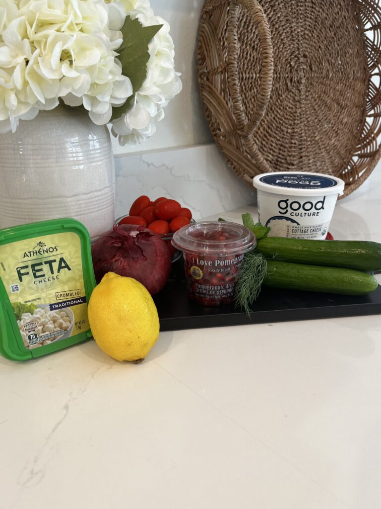 cottage cheese, pomegranate seeds, feta cheese, Persian cucumbers, red grape tomatoes, red onion, lemon, mint leaves, dill herb on the counter
