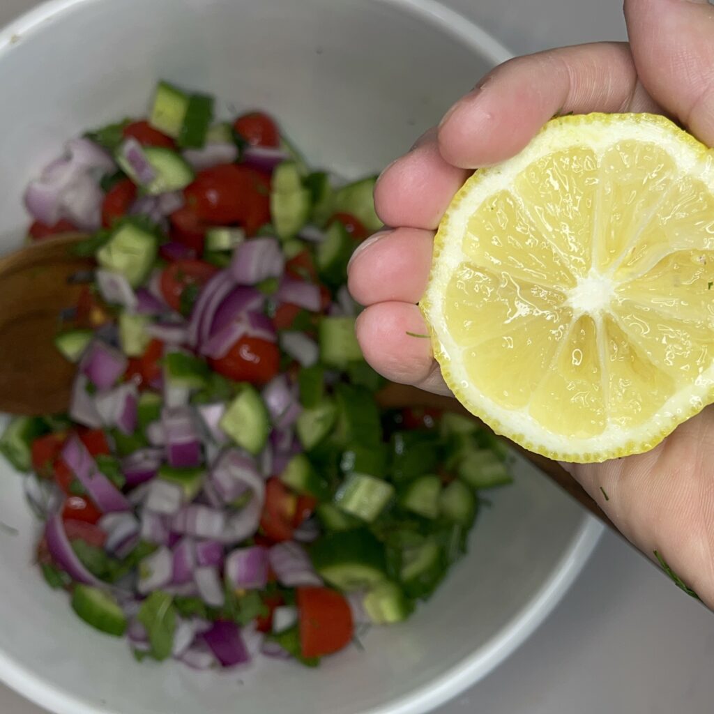 Diced cucumber, quartered tomatoes,  diced red onion in a large mixing bowl. And a  lemon about to be squeezed.
