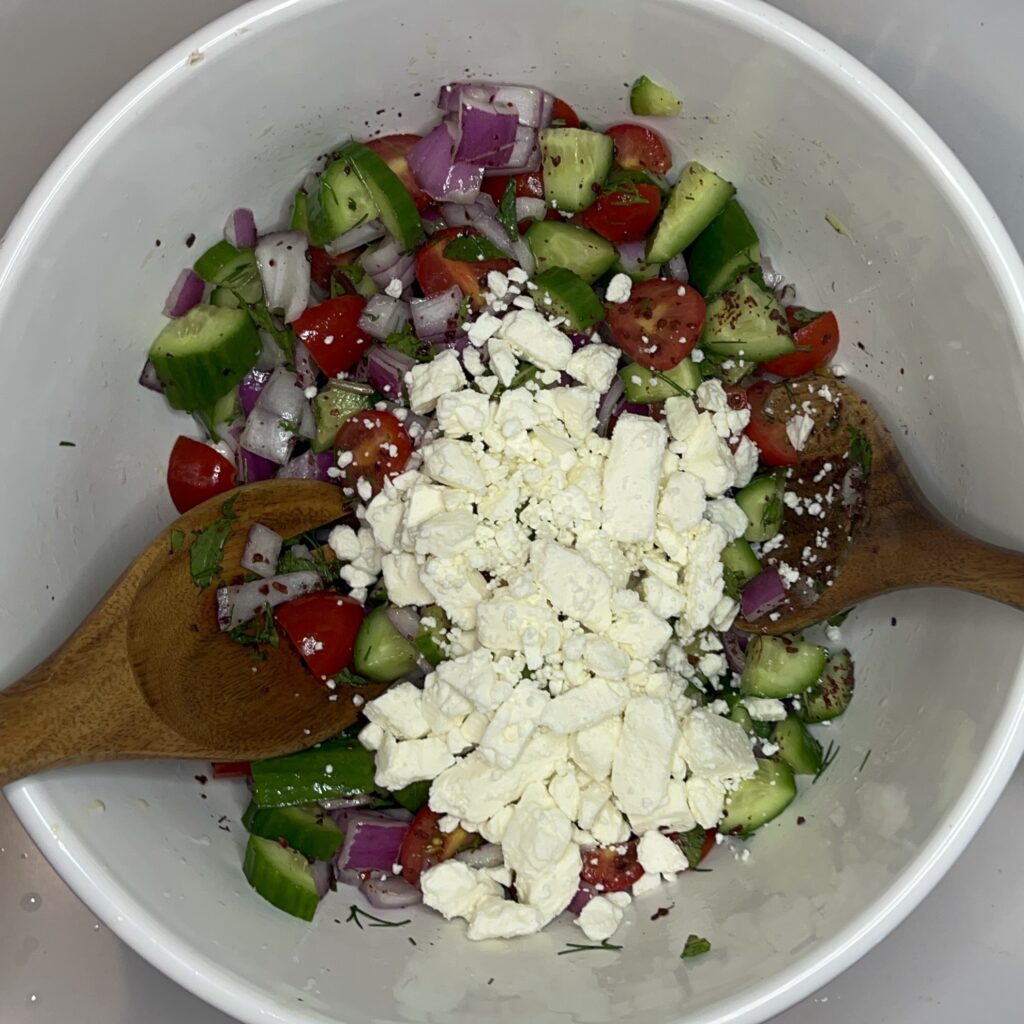 cucumber, tomatoes, red onion, mint, dill, pepper, lemon juice and feta in a large bowl being mixed.
