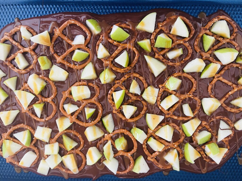 Melted chocolate on a baking sheet with green apple pieces pretzels and caramel sauce on a baking sheet