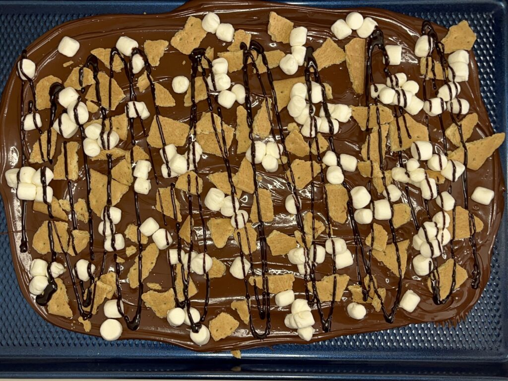 Melted chocolate on a baking sheet with mini marshmallows, graham crackers, and dark chocolate sauce on a baking sheet