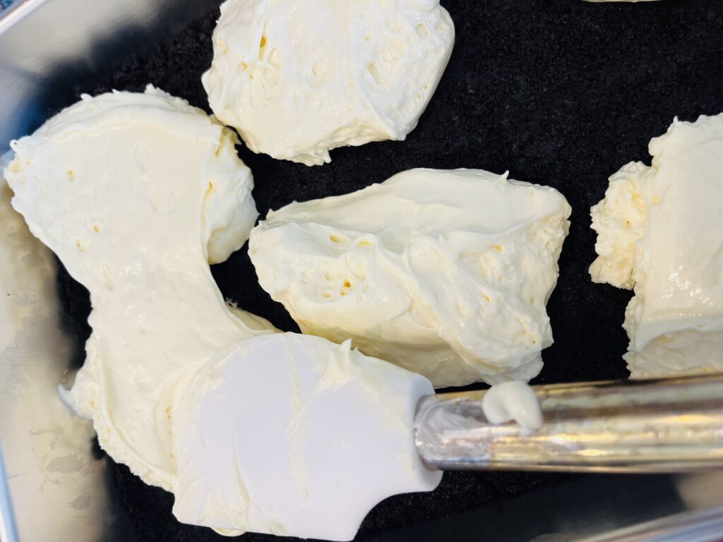 Using a spatula to spread out the cream cheese filling on top of the Oero cookie crust in a baking sheet pan