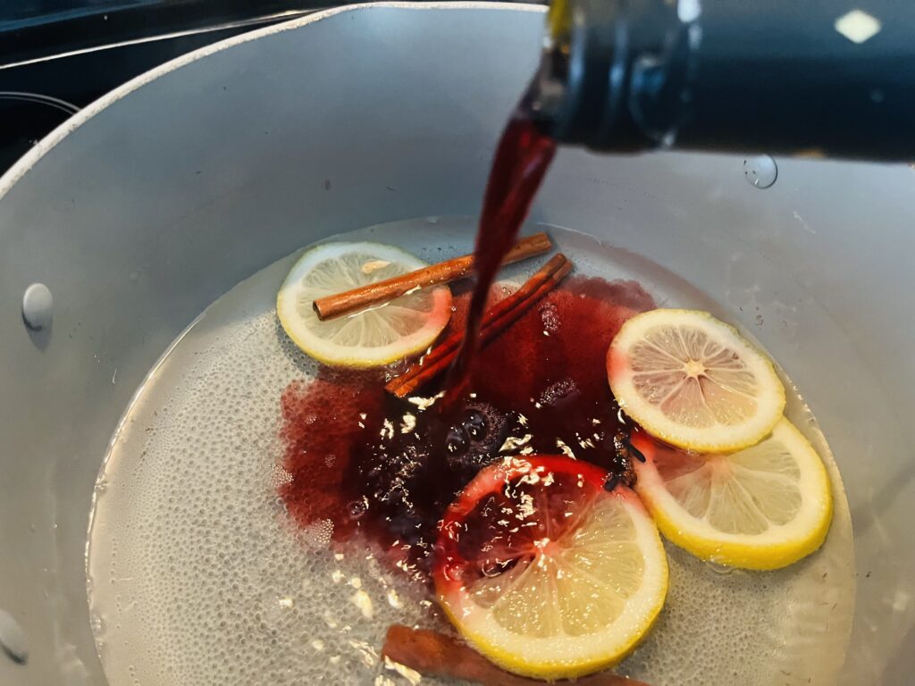 Pouring red wine into a pot on the stove with lemons, cinnamon sticks, cloves and sugar water.