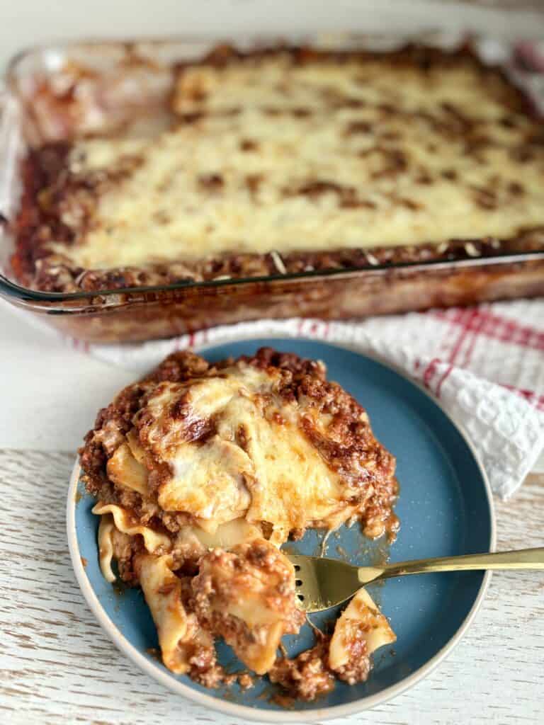 a slice of lasagna on a plate with a fork next to a casserole dish of lasagna