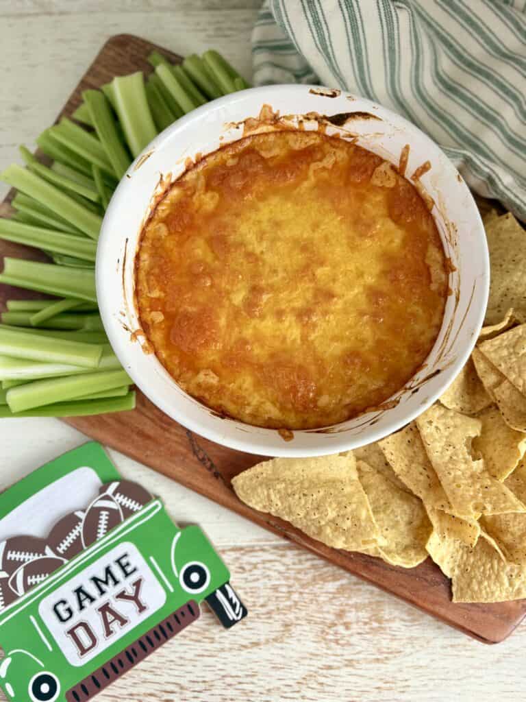 baked buffalo chicken dip in a white bowl on a board with celery and tortilla chips