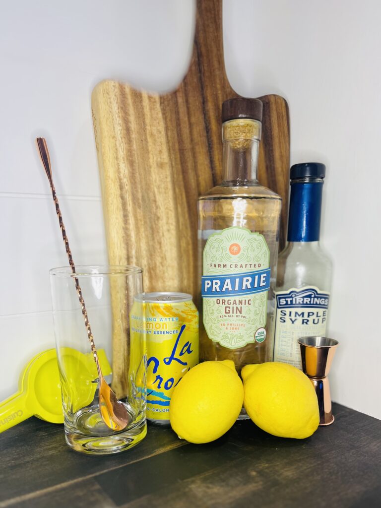 gin, simple syrup, lemons, a can of lemon flavored seltzer, a tall, glass, juicer, and spoon on a counter
