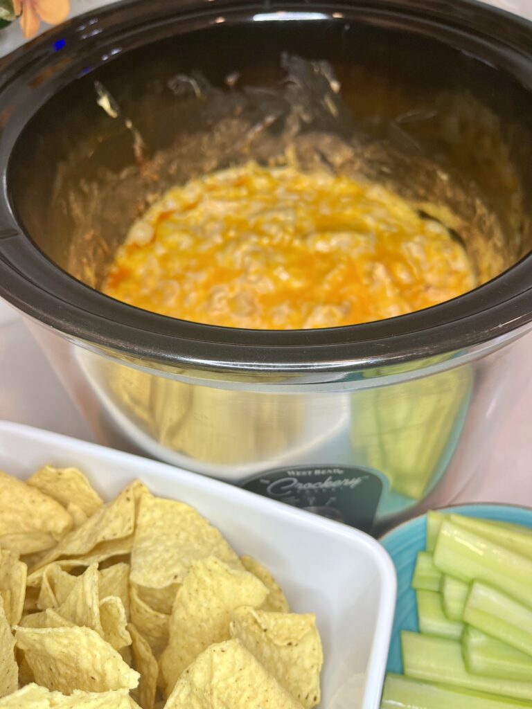 cooked dip in a slow cooker with a bowl of tortilla chips and a plate of celery