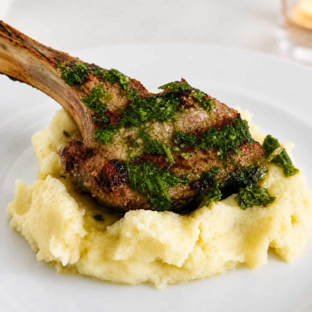 Lamb chop with chimichurri sauce served over creamy mashed potatoes on a white plate.