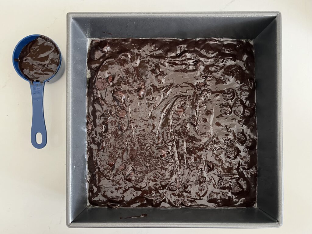 Dark chocolate brownie batter in a 9x9 baking pan with ¼ cup reserved on the side on the counter.