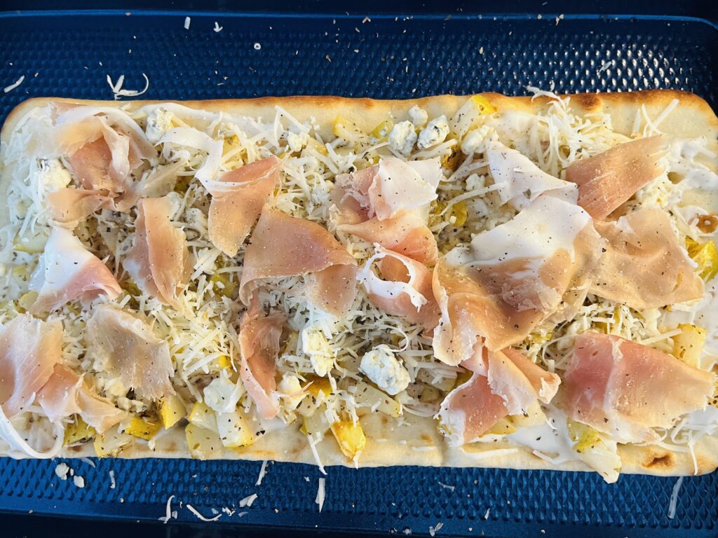 Flatbread topped with prosciutto slices, cheese, pears and seasonings on a baking sheet