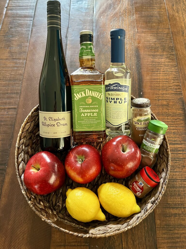 apples, sugar, cinnamon, nutmeg, allspice, lemons, simple syrup, apple-infused whiskey, allspice dram liqueur  in a basket on a table.