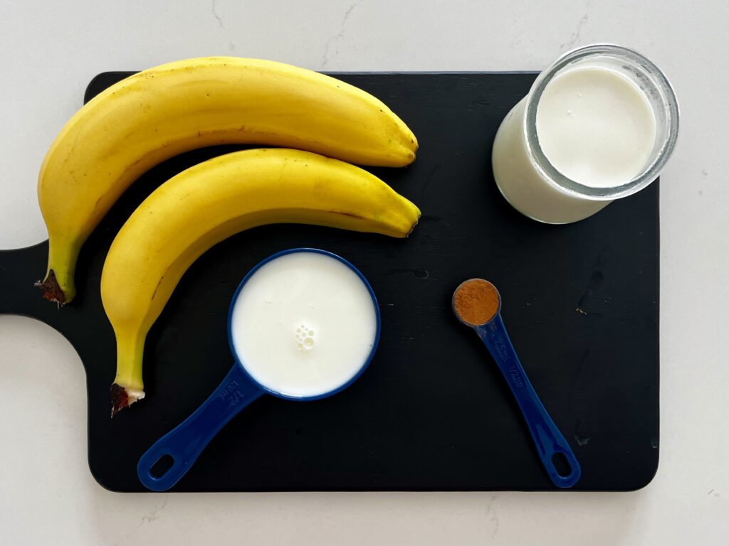 milk, two bananas, plain yogurt, cinnamon on a black cutting board.

