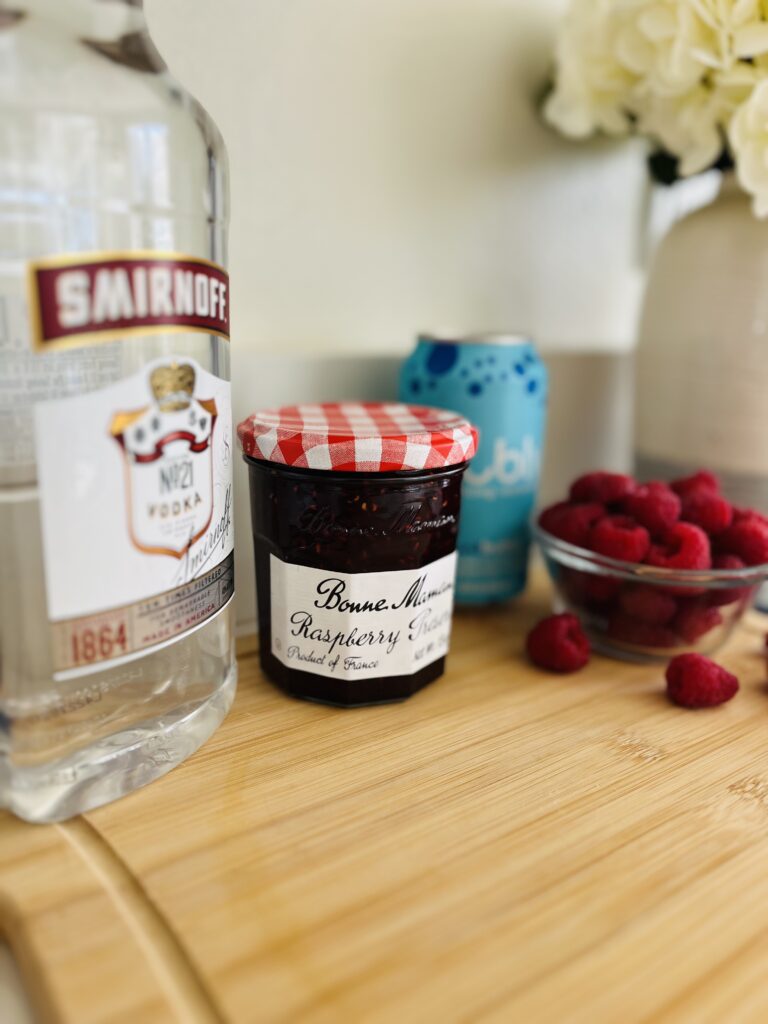 vodka, jam in jar, fresh raspberries, and club soda in a can on a cutting board on a counter.