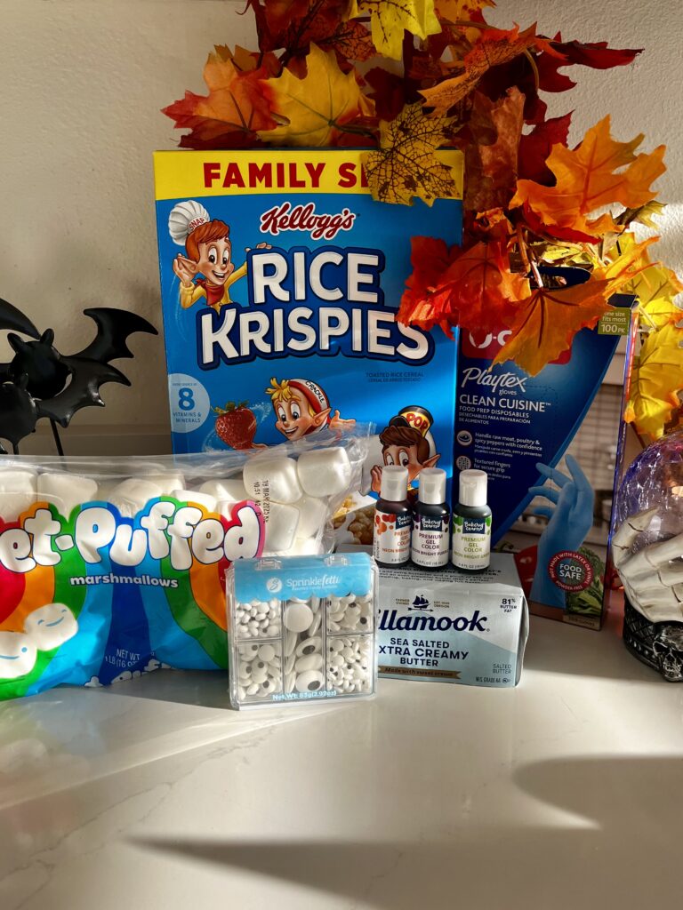 rice crispy cereal, neon food coloring, large marshmallows, food safe gloves, butter, sugar candy eyes and halloween decorations on the counter.