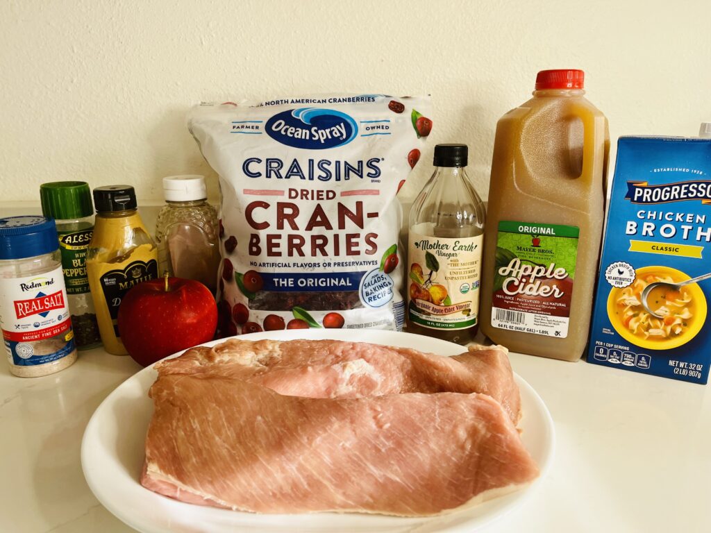 Pork loin fillets on a plate on the counter. cranberries, dijon mustard, apple cider, apple cider vinegar, apple, garlic, broth, honey, and salt and peeper on the counter.