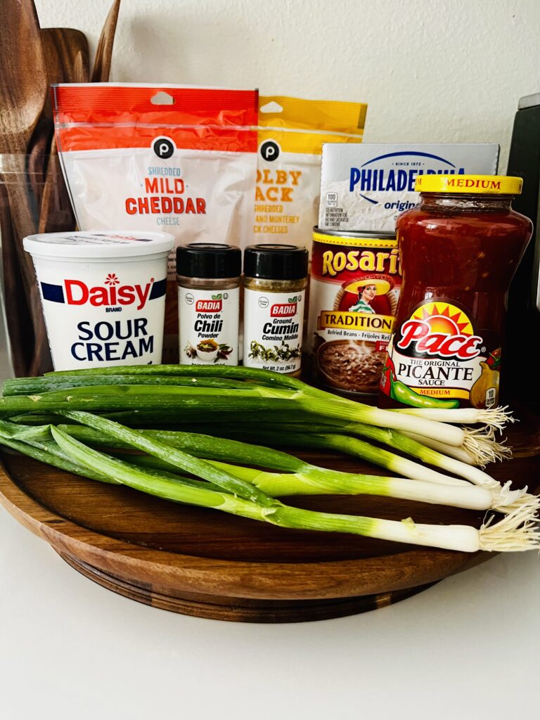 Cheeses, cream cheese, picante sauce, can of refried beans, spices, sour cream and green onions on a rotating tray on the counter.