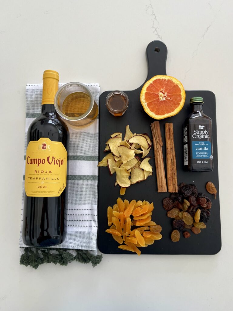 Bottle of red wine, glass of brandy, half an orange, jar of honey, apple chips, sliced apricots, raisons, vanilla extract, and cinnamon sticks on a black cutting board on the counter.