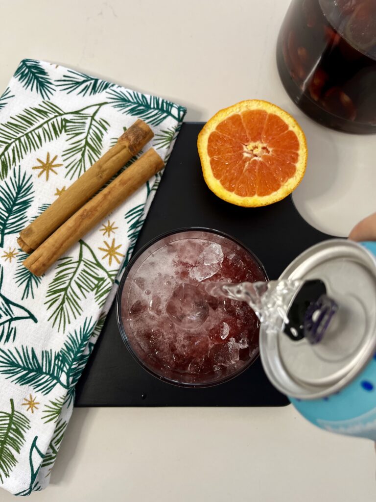 Pouring club soda into a glass with ice and sangria on a black cutting board on the counter.