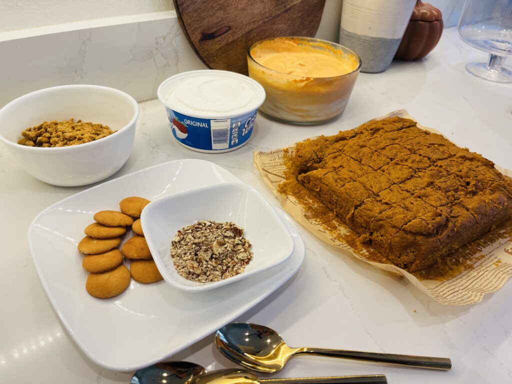 Cut up spice cake on parchment paper on the counter. Pumpkin cream cheese filling in a glass bowl, whip cream, crushed graham crackers I a bowl, crushed pecans in a small bowl and vanilla wafers on a plate- al on the counter.