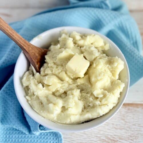 Creamy fluffy mashed potatoes in a white bowl with a wooden spoon, served on a rustic wooden table with a blue cloth napkin, perfect for comfort food and holiday dinners.