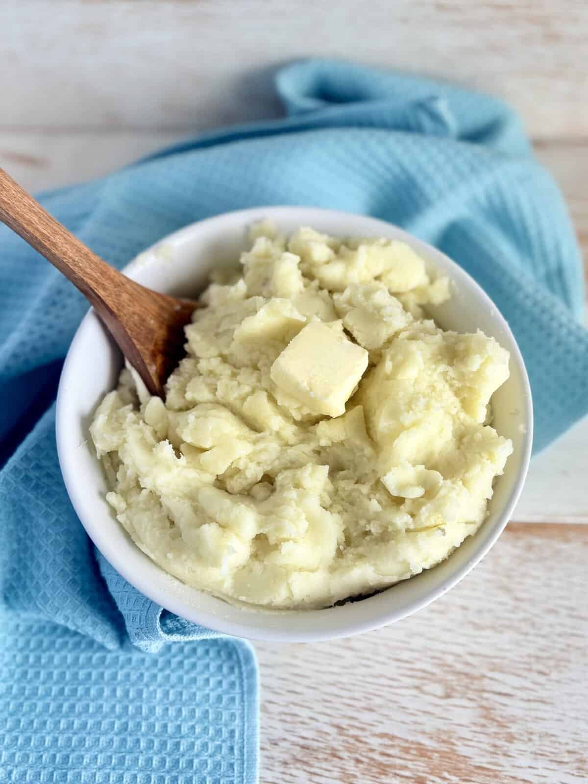 Creamy fluffy mashed potatoes in a white bowl with a wooden spoon, served on a rustic wooden table with a blue cloth napkin, perfect for comfort food and holiday dinners.
