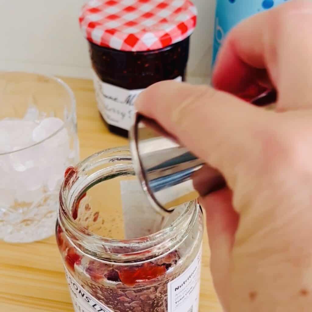 Pouring vodka inside of jam that's been left over for remaining in a jam jar with strawberry jam and a glass full of ice in the background