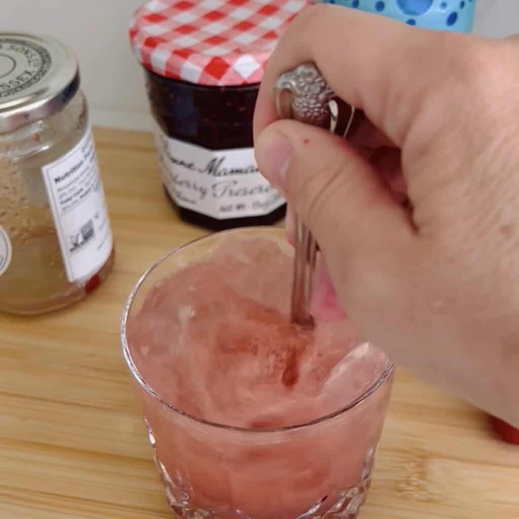 Using a spoon to stir the thick jam, citrus, juice, and soda altogether and a clear glass full of ice