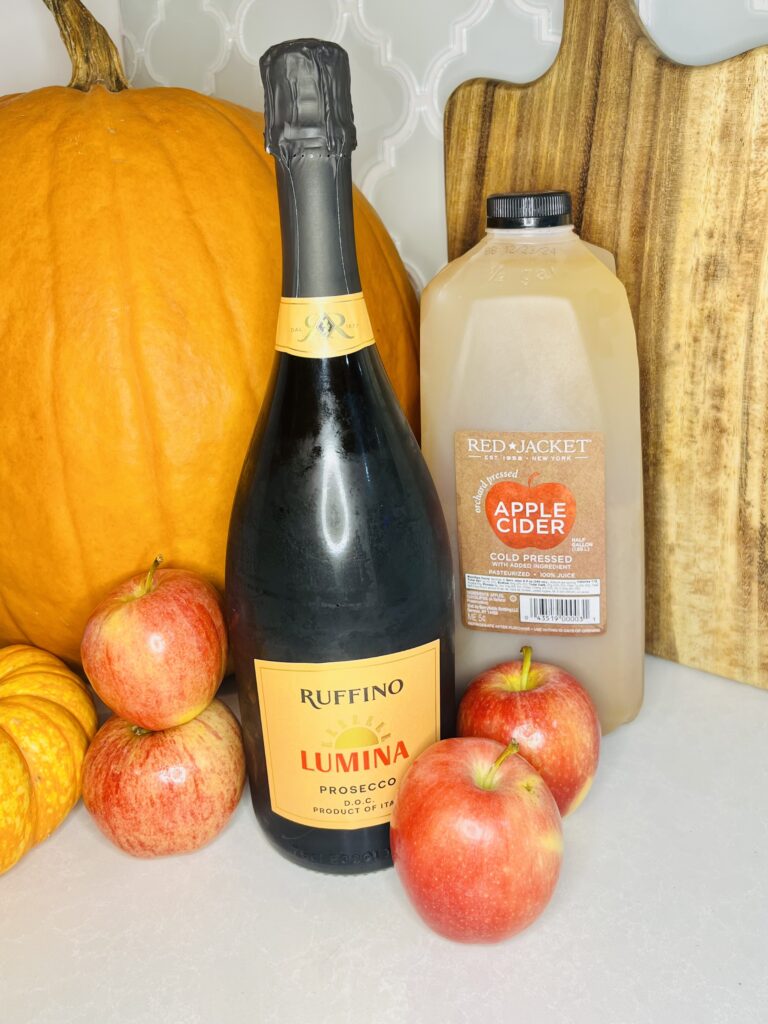 a bottle of Prosecco with apples, apple cider, and a pumpkin on a counter