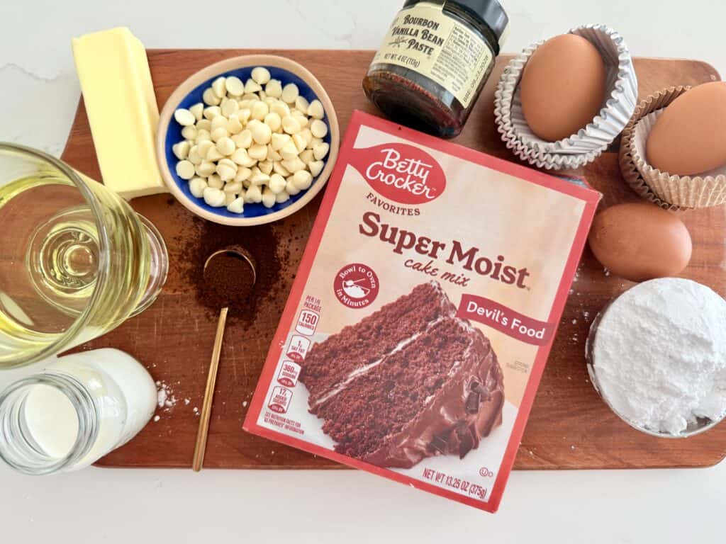  a box of devil's food cake mix, white chocolate chips in a blue bowl. vanilla bean paste. 3 eggs, vegetable oil, instant esspresso powder, heavy cream, unsalted butter, powdered sugar all on a brown cutting board.