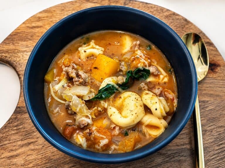 Hearty autumn tortellini soup with vegetables and ground meat in a dark bowl on a wooden tray.