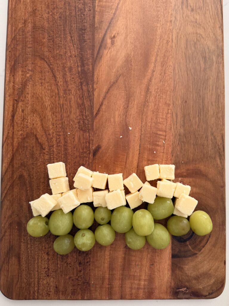 Green grapes topped with cubes of white cheddar cheese on a brown board on the counter.