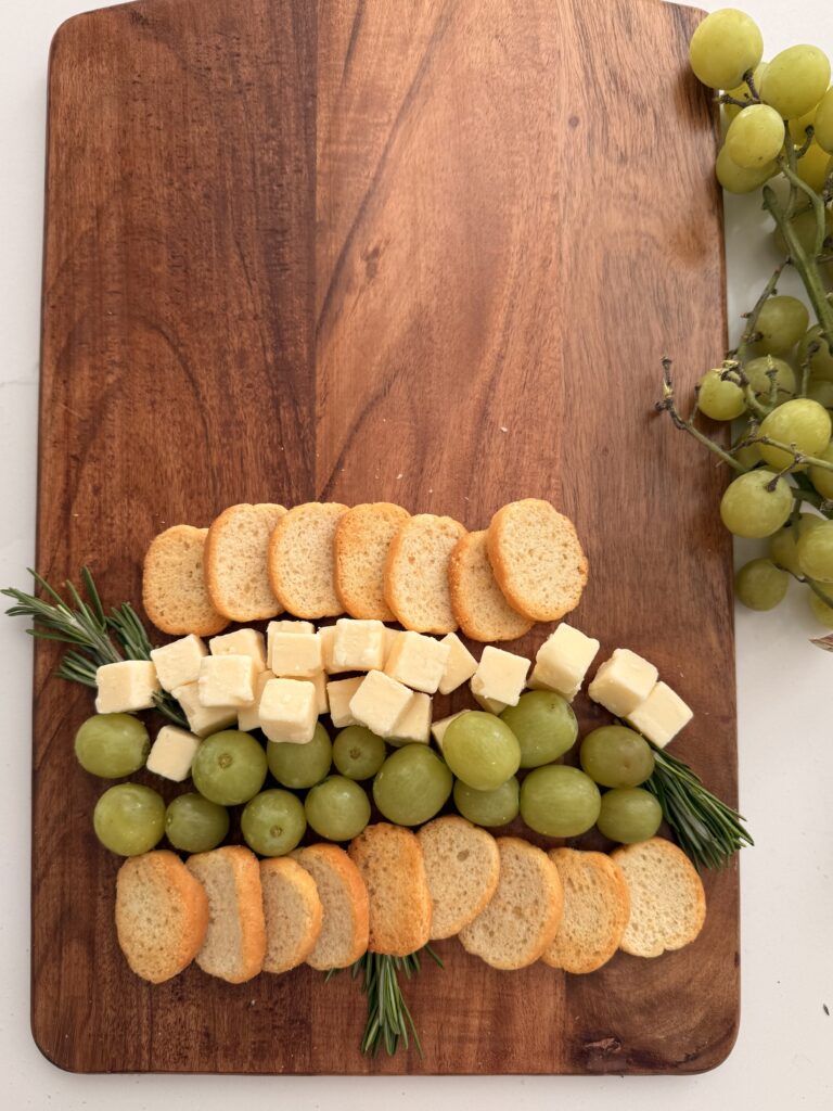 Crackers, green grapes topped with cubes of white cheddar cheese and then topped with more crackers on a brown board on the counter. Sprigs of rosemary as tree trunk and decoration between cheese and grapes on a brown board on the counter.