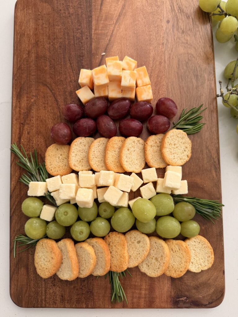 Crackers, green grapes topped with cubes of white cheddar cheese and then topped with more crackers and then red grape and then more cubes orange cheese on a brown board on the counter in a Christmas tree shape. Sprigs of rosemary as tree trunk and decoration between cheese and grapes on a brown board on the counter.