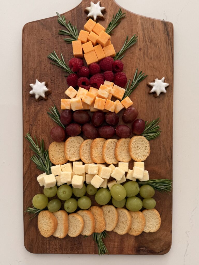 Crackers, green grapes topped with cubes of white cheddar cheese and then topped with more crackers and then red grape and then more cubes of orange cheese, next topped with red raspberries and then more cubed cheese in a Christmas Tree shape topped with a star shaped cookie on a brown board on the counter. Sprigs of rosemary as tree trunk and decoration between cheese and grapes on a brown board on the counter.