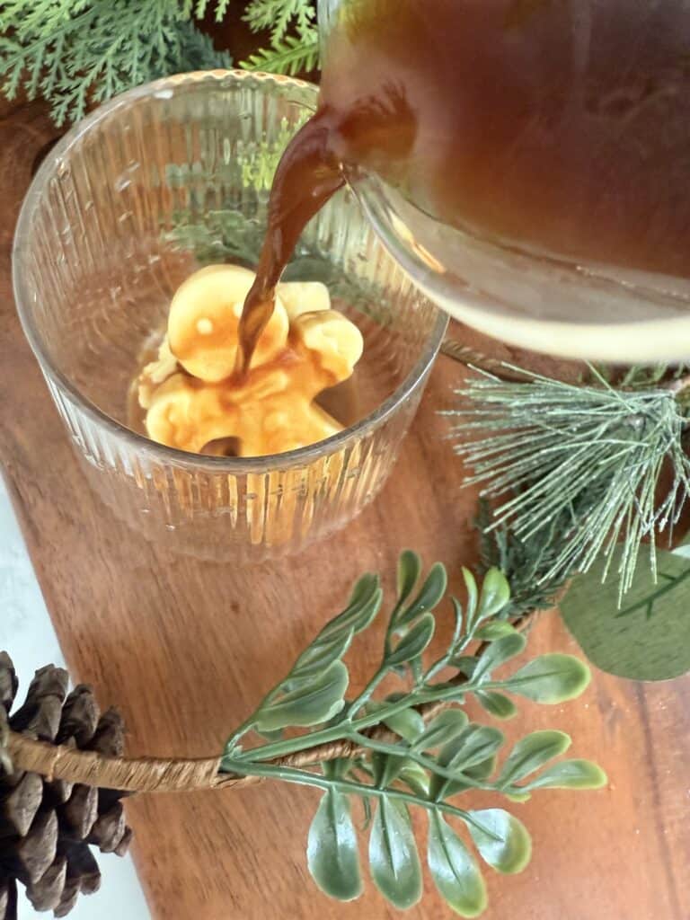 Creamy coffee being poured into a glass mug with eggnog festive gingerbread man ice cube, surrounded by festive greenery and pinecone, perfect for holiday drinks and cozy winter gatherings.