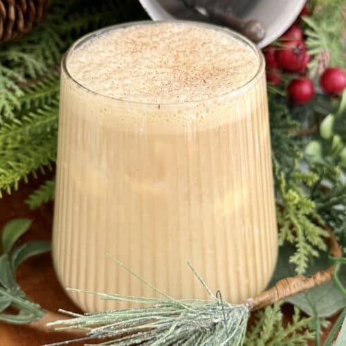 Creamy eggnog in a clear glass, topped with nutmeg, surrounded by holiday greenery and rustic decorations for a festive seasonal beverage.