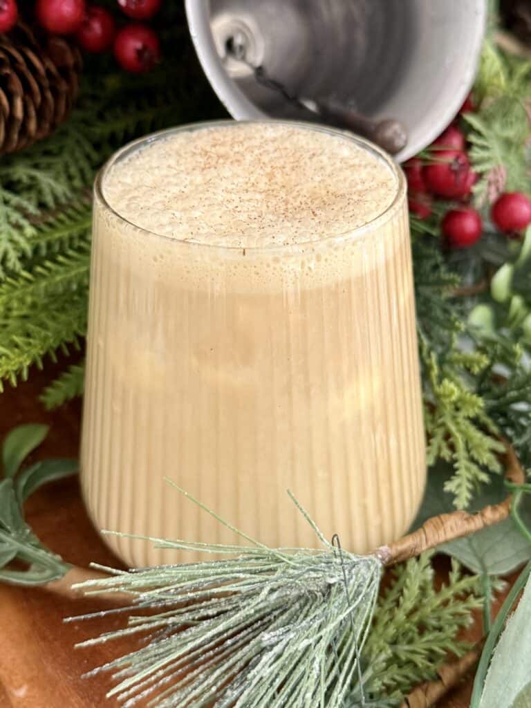 Creamy eggnog in a clear glass, topped with nutmeg, surrounded by holiday greenery and rustic decorations for a festive seasonal beverage.