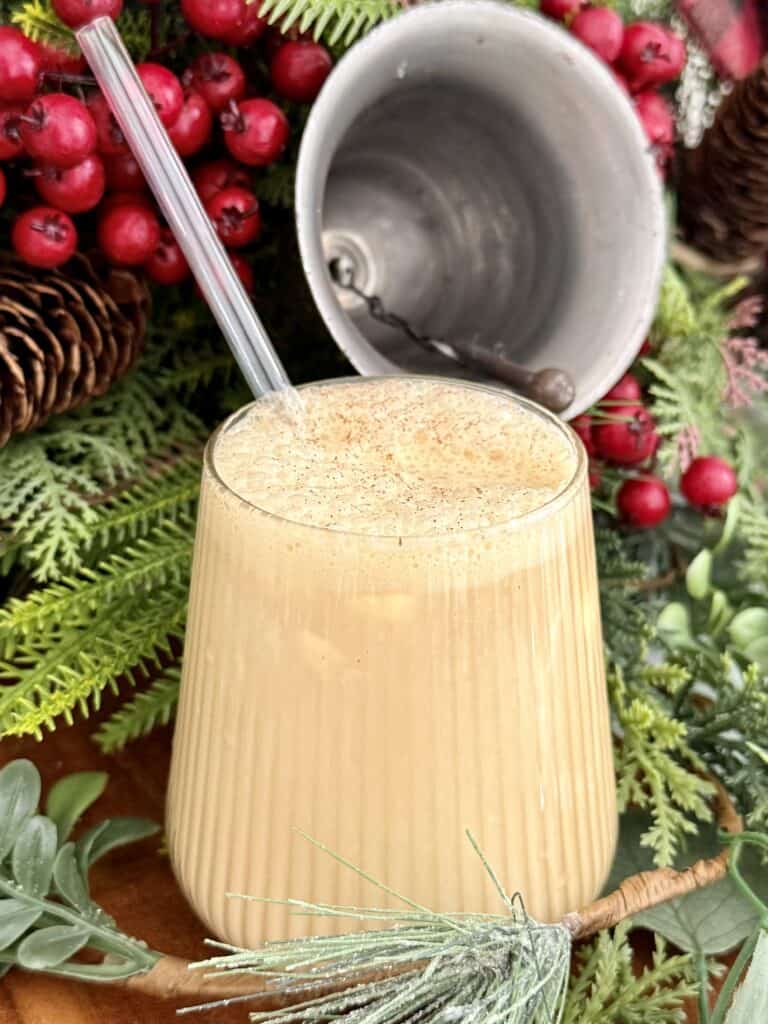 Rich and creamy eggnog in a glass with a straw, surrounded by festive holiday decorations and greenery, perfect for Christmas celebrations.