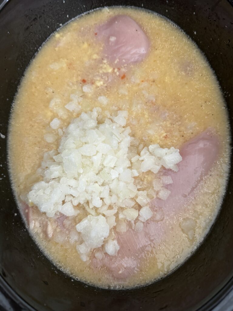 Chopped onions on top of chicken and liquid ingredients in the slow cooker on the counter.