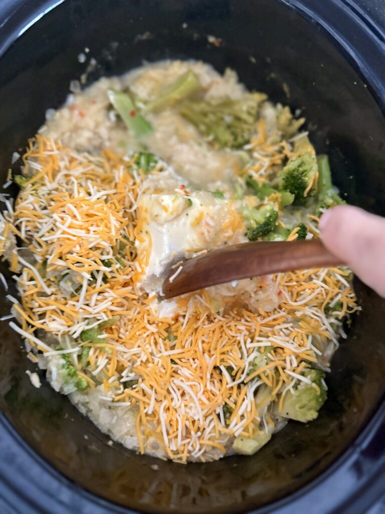 Stirring with a wooden spoon the broth, rice, broccoli, colby cheese, cream cheese, italian dressing, and soup in the slow cooker on the counter.